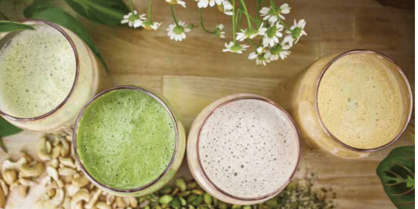 Top-down view of four colorful smoothies in glasses on a wooden table, surrounded by nuts and herbs with white flowers above.
