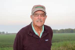 Portrait of an older man with glasses and a mustache standing in a green farmland landscape, wearing a beige cap and a dark zippered sweater over a teal collared shirt