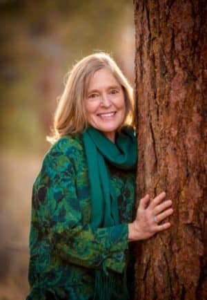 Older woman with a warm smile stands beside a tree in a forest, wearing a green scarf and patterned teal-green sweater.