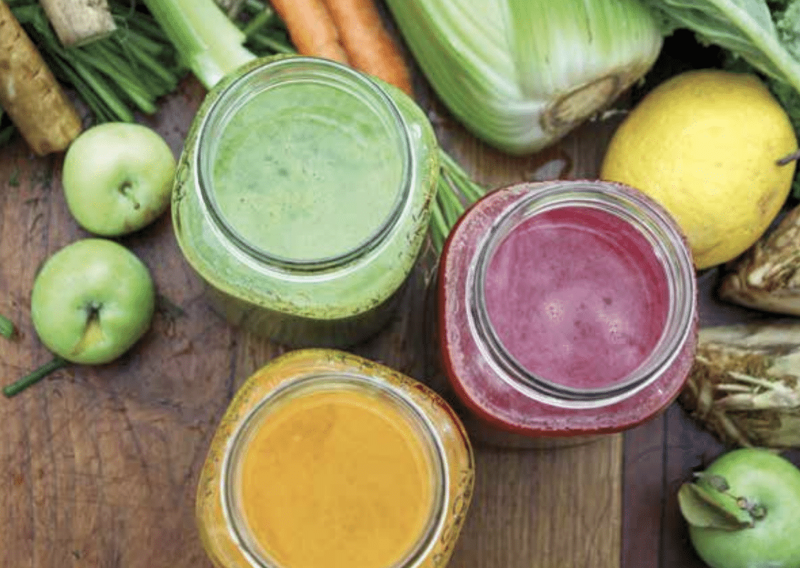Three glass jars of colorful smoothies (green, pink, orange) on a wooden table with fresh produce around them.