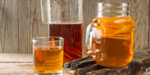 Mason jar and glass of amber tea beside cinnamon sticks on a wooden table surface.