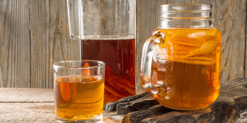 Mason jar and glass of amber tea beside cinnamon sticks on a wooden table surface.