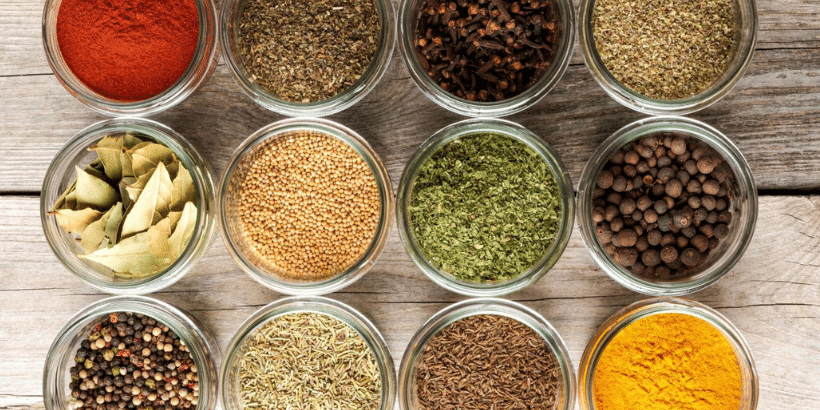 Top-down view of twelve glass spice jars arranged on a wooden table, showing red paprika, seeds, and dried herbs in different shades.