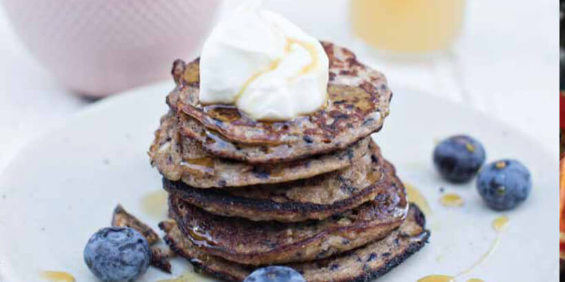 A stack of five pancakes on a plat topped with whipped cream and blueberries