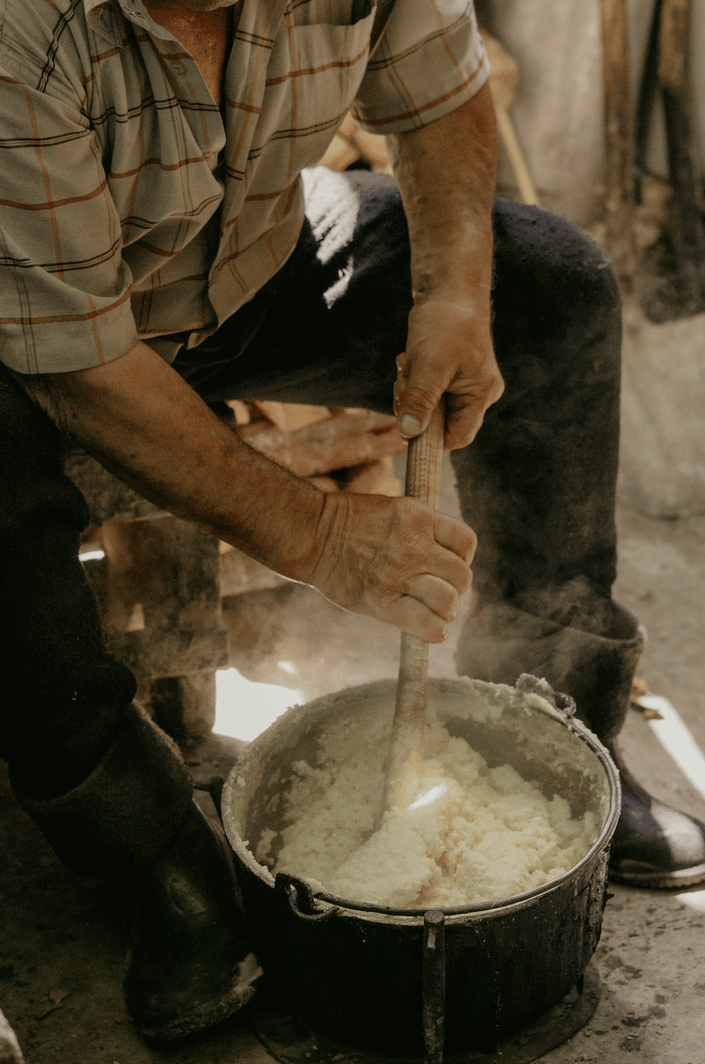 Elderly man stirring a large pot of mashed potatoes with a wooden spoon outdoors.
