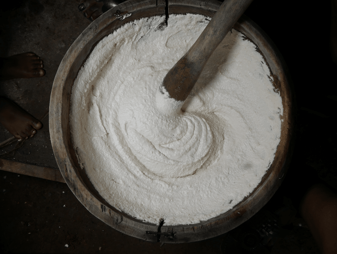 Large wooden tub filled with a white, powdery paste being stirred with a wooden paddle; bare feet visible nearby.