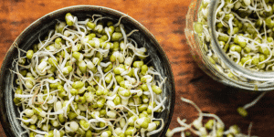 Bowl of fresh mung bean sprouts on a wooden table with more sprouts in a jar nearby.