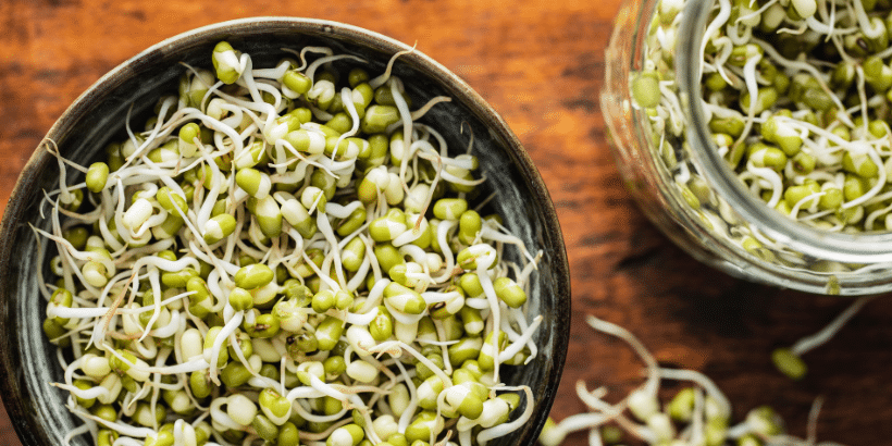 Bowl of fresh mung bean sprouts on a wooden table with more sprouts in a jar nearby.