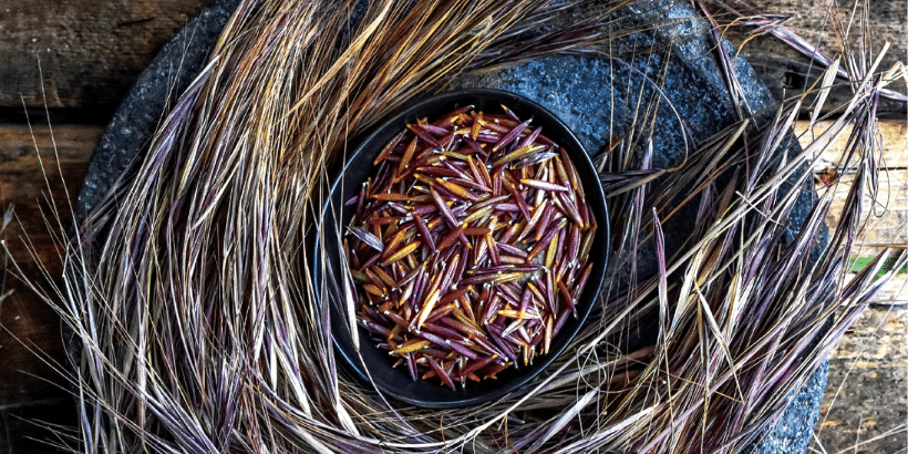 Black bowl filled with purple-red rice grains, nestled in a circular bed of dried straw on a wooden surface.