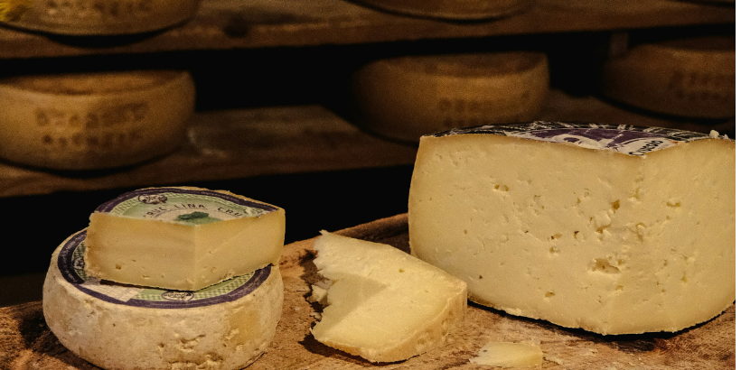 Round wheel of cheese with a wedge cut, on a wooden board, with more cheese wheels in the background.