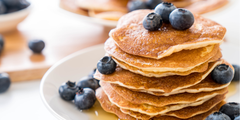Stack of golden pancakes topped with fresh blueberries on a white plate, drizzled with syrup.