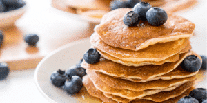 Stack of golden pancakes topped with fresh blueberries on a white plate, drizzled with syrup.