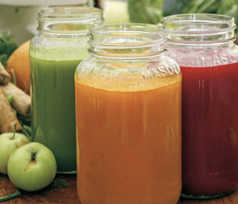 Three mason jar bottles of fresh juice in green, orange, and red, set on a wooden table with apples nearby.