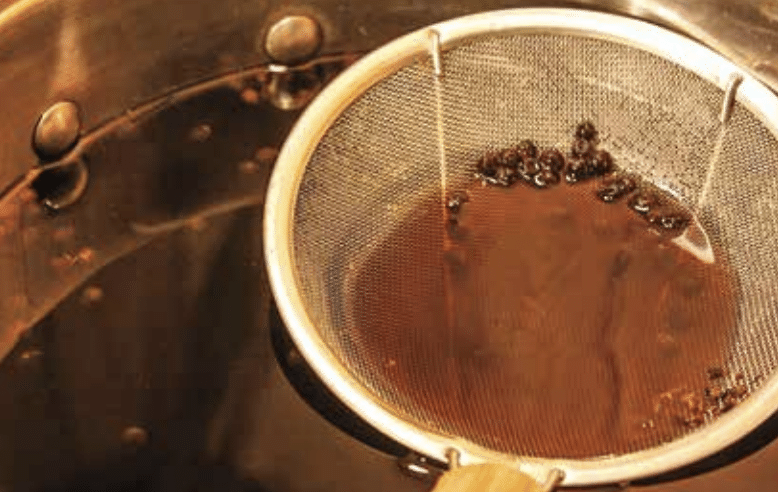 Close-up of a metal coffee filter draining dark brown coffee, with coffee beans resting in the liquid.