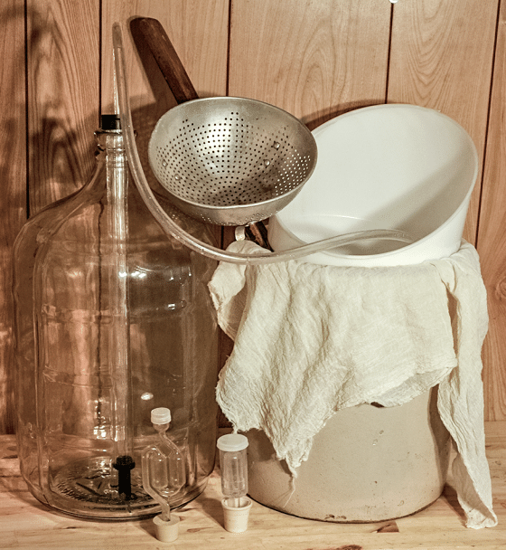 Home brewing setup with a large glass carboy, tubing, a metal strainer, and a white bucket covered with a towel.