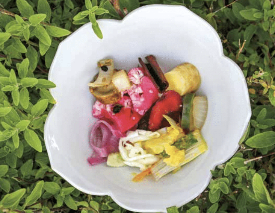 White scalloped bowl on green plants, filled with colorful pickled vegetables and edible flowers, creating a vibrant side dish presentation.