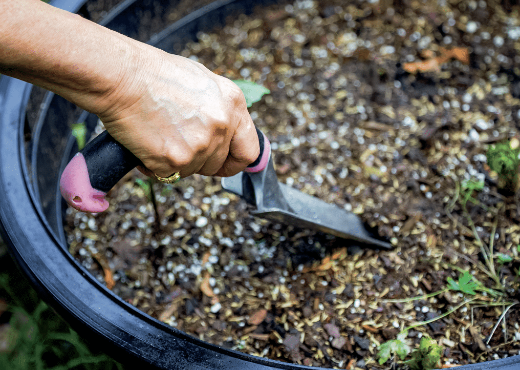 Person's hand with a pink-handled garden trowel digging into soil in a raised garden bed.